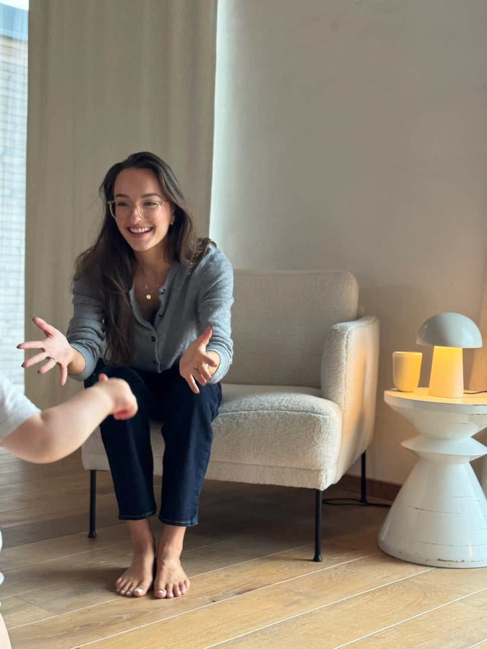 Person sitzt lächelnd im Wohnzimmer und spielt mit einem Kleinkind — warmer Familienmoment in natürlicher Glowbug-Atmosphäre