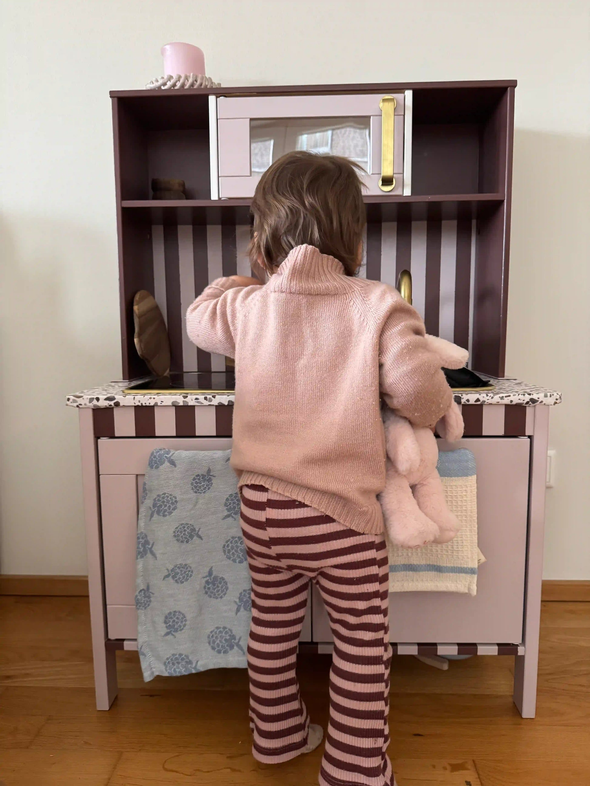 Kleinkind spielt an einer Kinderküche und hält ein Kuscheltier — warmer Familienmoment im Alltag mit Glowbug-Gefühl von Geborgenheit